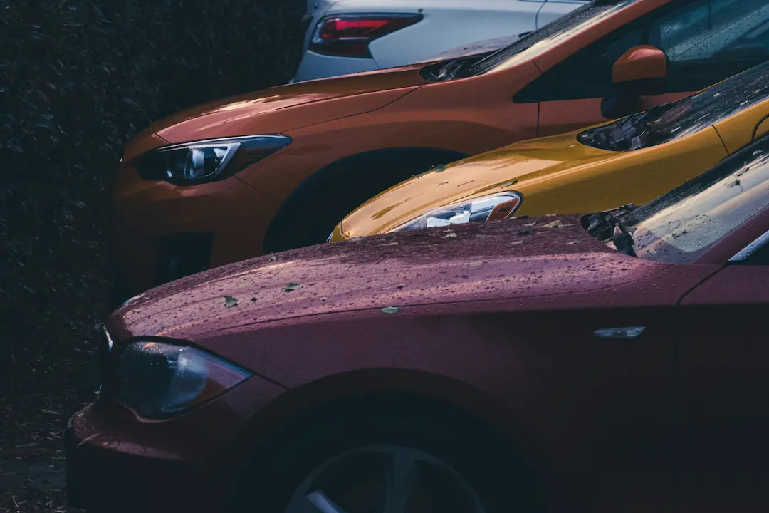 Trzy zaparkowane samochody – bordowy, żółty i pomarańczowy – częściowo na siebie zachodzą na zacienionym parkingu; ich pokryte kroplami deszczu maski i reflektory tworzą nastrojową, filmową scenerię.