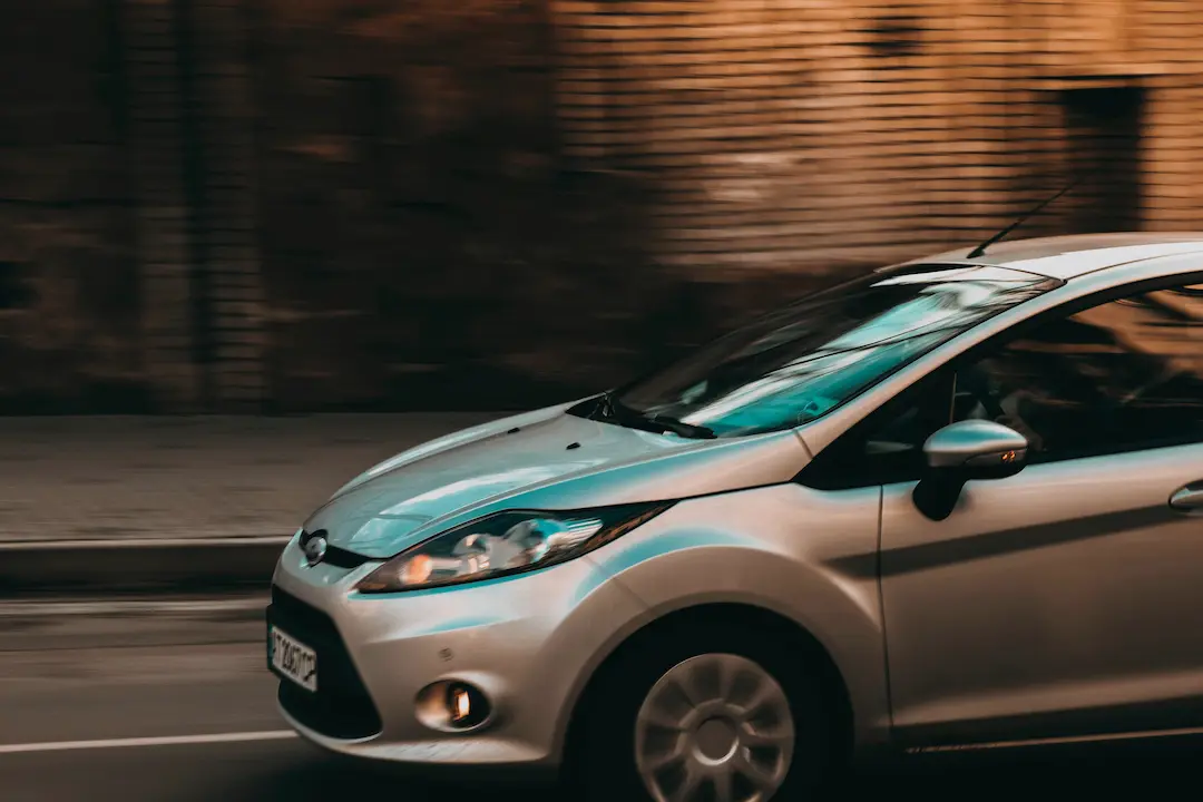 Srebrny, kompaktowy hatchback pędzi miejską ulicą o zmierzchu; rozmyte tło z ceglaną zabudową oraz refleksy światła oddają wrażenie ruchu i nadają całości smukły, pełen energii, miejski klimat.